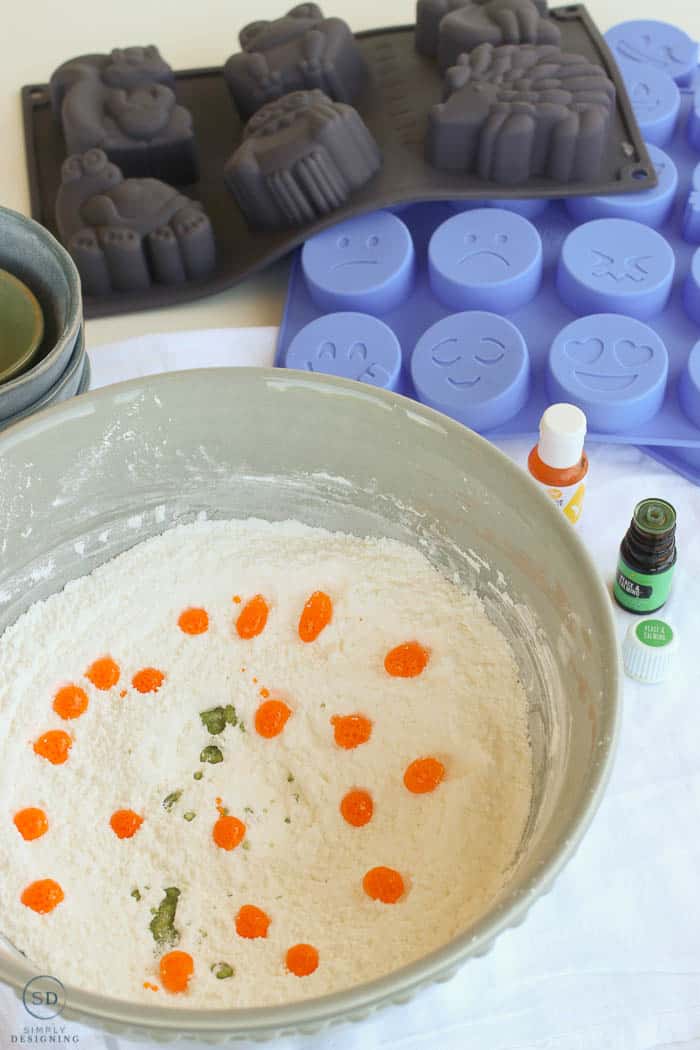 Vertical image showing the food coloring, essential oil, and dry ingreidents in a large blue bowl. Making kids bath bombs with emoji molds in the background. 