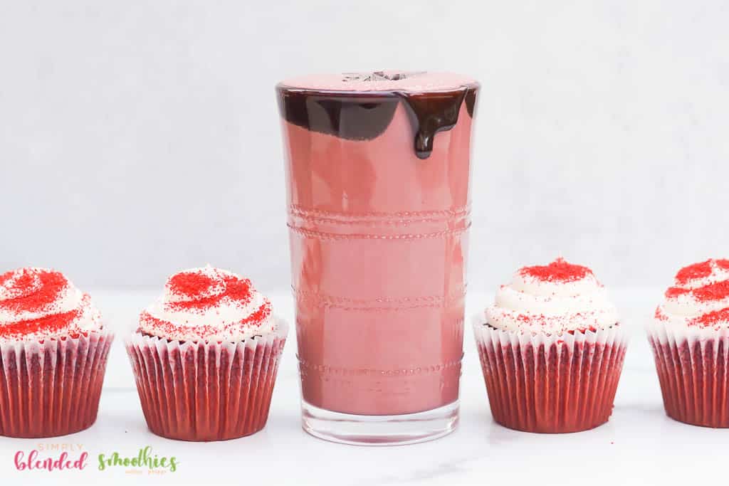 red velvet shake with chocolate syrup dripping down the side chocolate flakes and red velvet cupcakes on the side