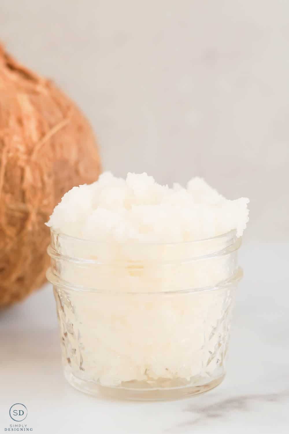 vertical photo of coconut body scrub in a small glass mason jar with a partial view of a coconut in the background