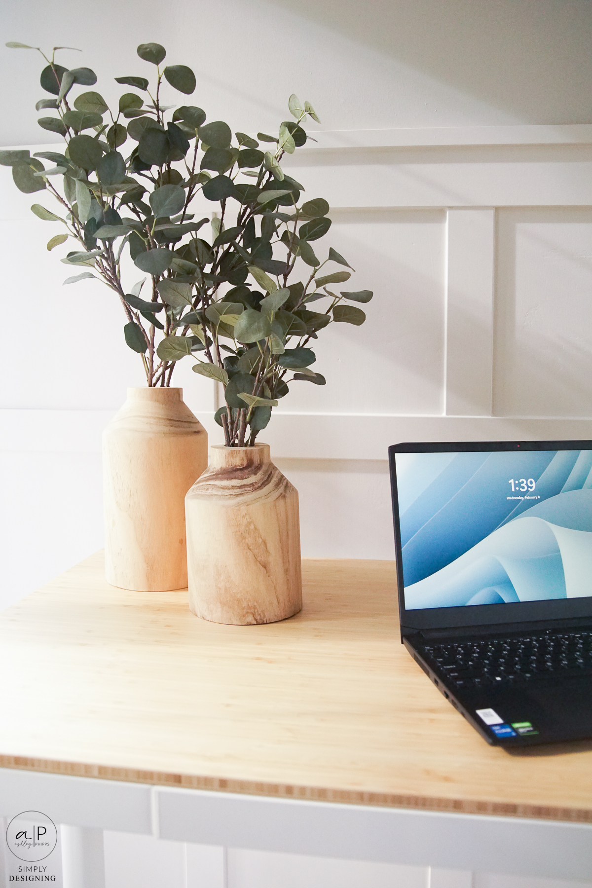 wood vases with fake greenery in it on a light wood top standing desk with half a black laptop in the frame